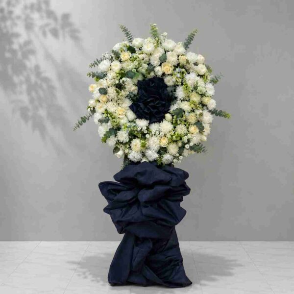Kệ hoa trắng 01 với vòng hoa chia buồn tông trắng kết hợp điểm nhấn nơ xanh đen sang trọng, thể hiện sự thành kính và tiếc thương