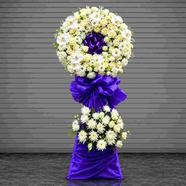 Kệ chia buồn tiễn biệt 420 với vòng hoa trắng cao cấp kết hợp hoa lan hồ điệp trắng, hoa hồng trắng và nơ tím nổi bật, mang vẻ đẹp trang nghiêm và sang trọng.