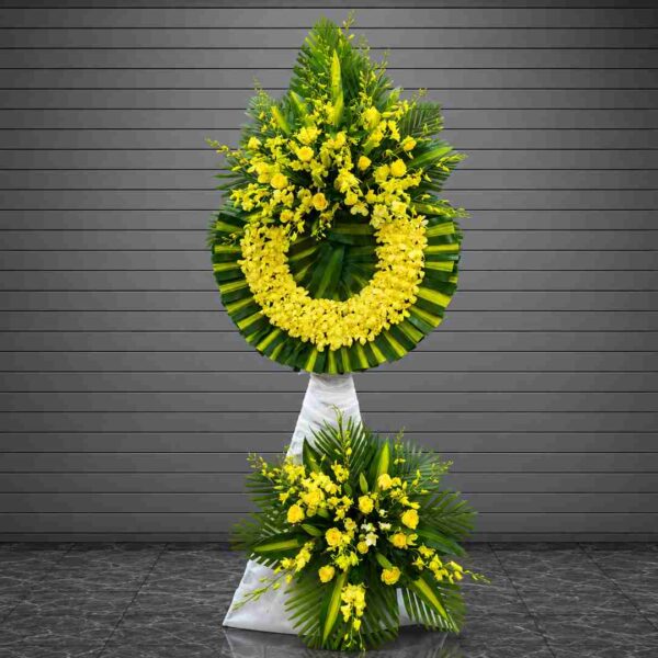 Kệ hoa chia buồn 115 với vòng hoa lan vàng và hoa hồng vàng nổi bật, kết hợp lá xanh nghệ thuật, mang thông điệp tưởng nhớ và chia buồn trang trọng.