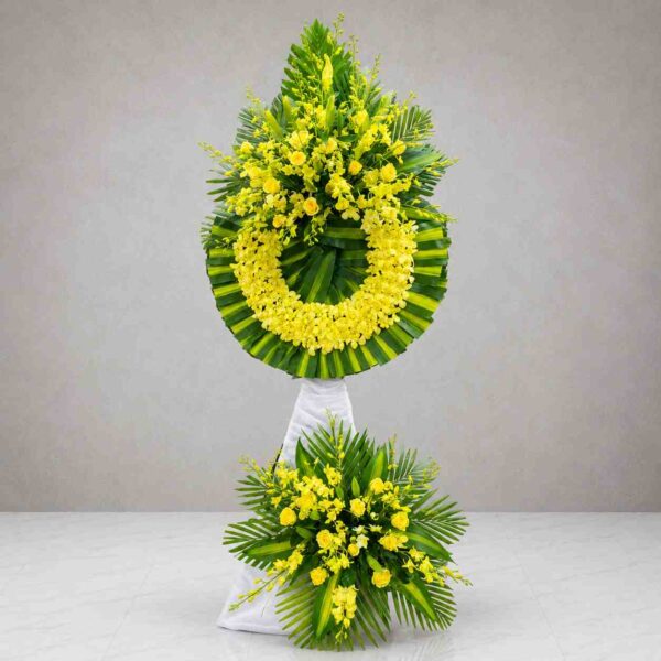 Kệ hoa chia buồn 115 với vòng hoa lan vàng và hoa hồng vàng nổi bật, kết hợp lá xanh nghệ thuật, mang thông điệp tưởng nhớ và chia buồn trang trọng.