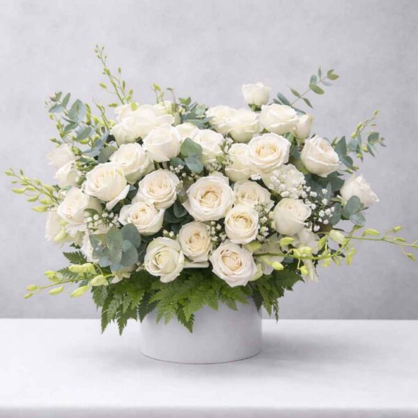 Giỏ hoa viếng chia xa màu trắng sang trọng với hoa hồng trắng, hoa baby và lá xanh đặt trên ghế gỗ nền cửa cuốn xám