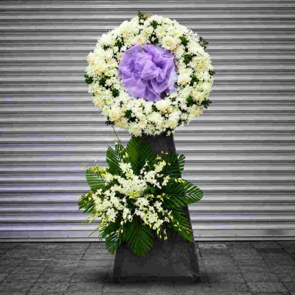 Kệ hoa chia buồn 501 với vòng hoa tròn màu trắng kết hợp nơ tím trang nhã, điểm nhấn hoa lan trắng và lá cau xanh, thể hiện lòng tiếc thương và sự kính trọng sâu sắc.