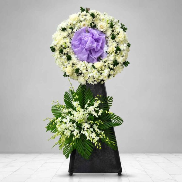 Kệ hoa chia buồn 501 với vòng hoa tròn màu trắng kết hợp nơ tím trang nhã, điểm nhấn hoa lan trắng và lá cau xanh, thể hiện lòng tiếc thương và sự kính trọng sâu sắc.