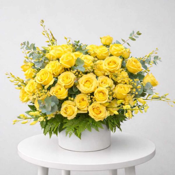 Giỏ hoa viếng màu vàng sang trọng với hoa hồng vàng, lá xanh, thiết kế tinh tế đặt trên bàn tròn trắng nền xám nhạt